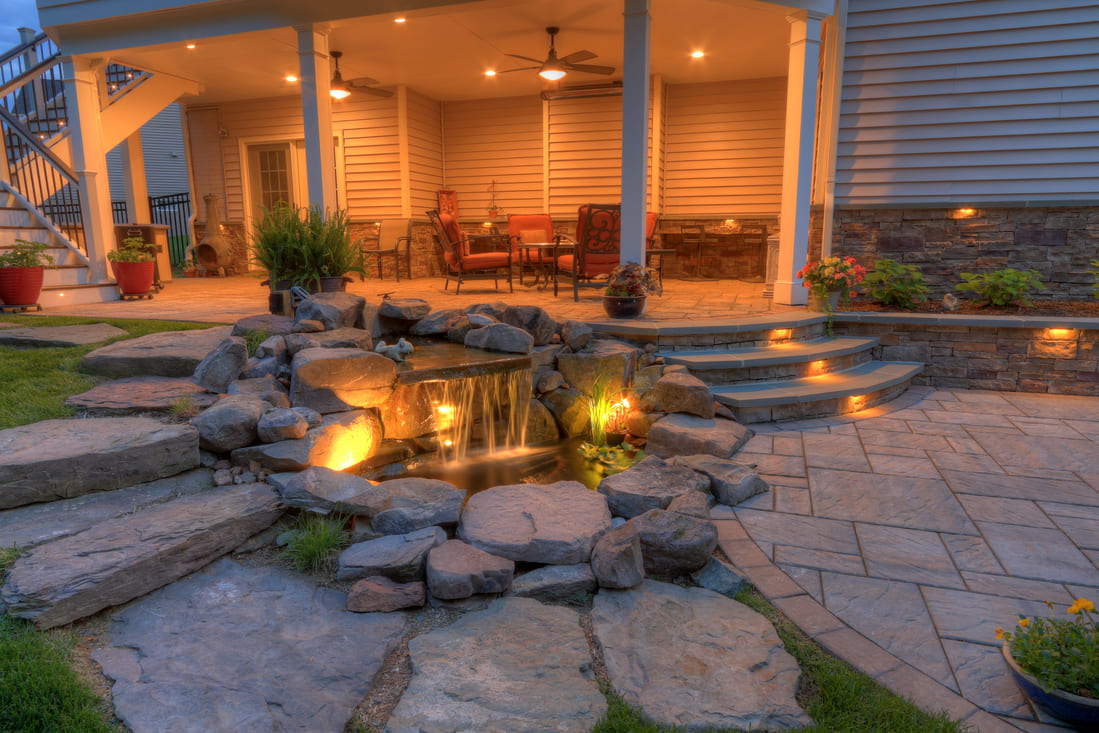 Stone patio waterfall next to pathway at dusk with custom lighting 